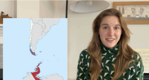 A woman with long, light brown hair and a green patterned sweater sits indoors. Beside her is a map showing southern South America with highlighted regions in red.
