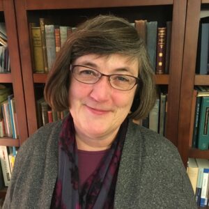 A woman with short gray hair and glasses smiles in front of a bookshelf filled with books. She is wearing a purple top, a patterned scarf, and a gray cardigan.
