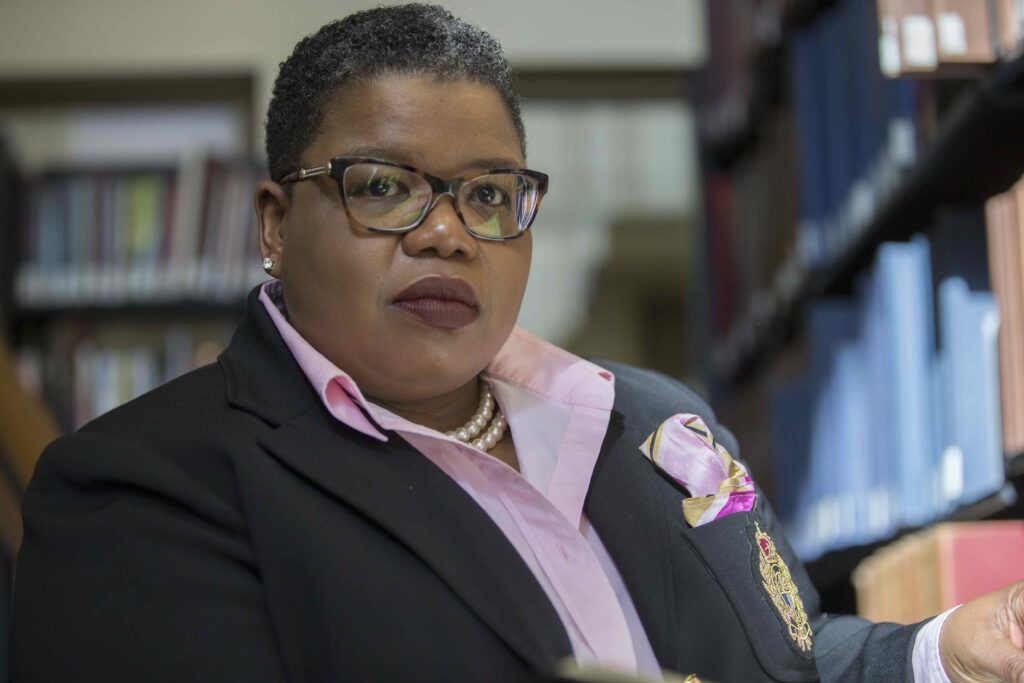 A person wearing glasses, a black blazer, and a pink shirt sits in a library, looking confidently at the camera. Bookshelves are visible in the background.