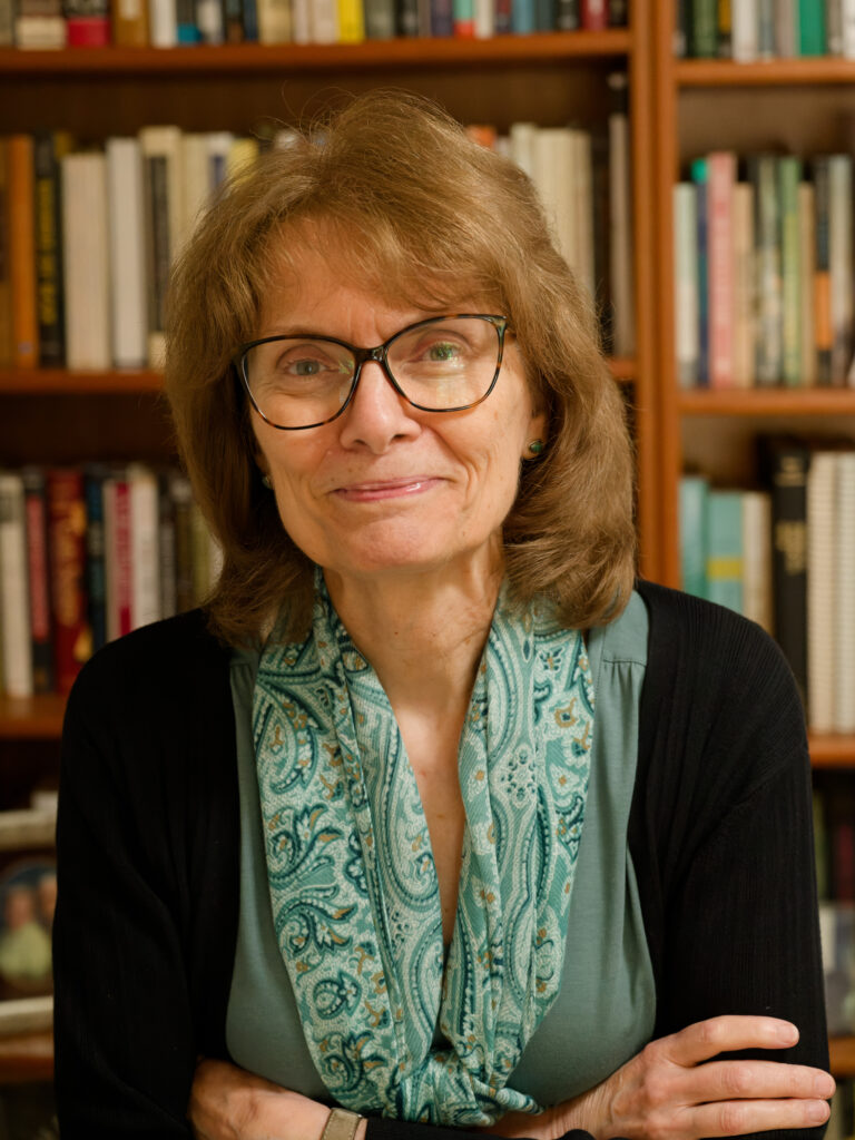 A woman with glasses and shoulder-length hair smiles softly, wearing a green top and patterned scarf, standing in front of bookshelves filled with books.