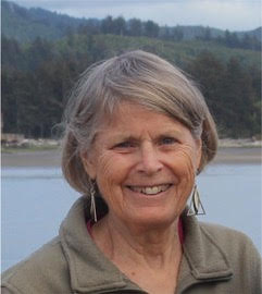 An older woman with short gray hair and earrings smiles outdoors near a body of water, with trees and hills in the background.