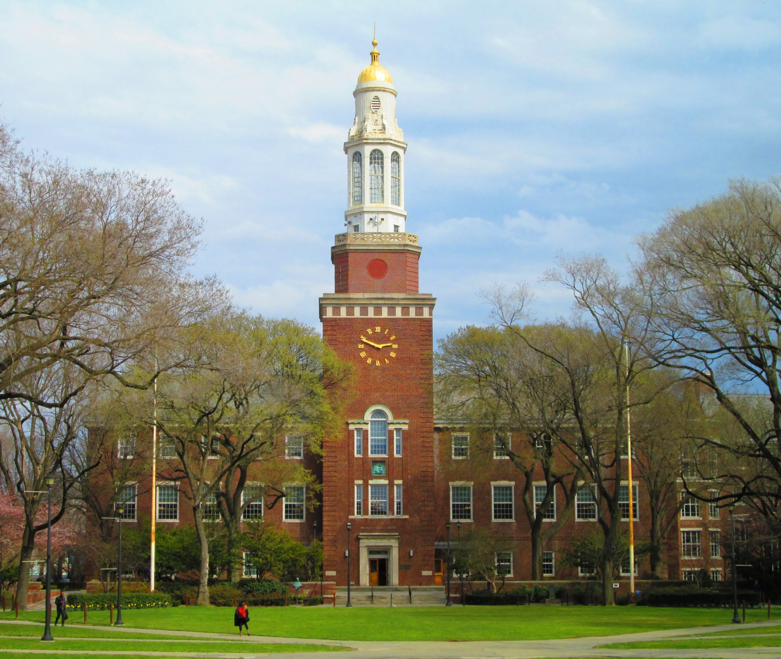A large brick building with a tall white clock tower and gold dome, surrounded by trees. People are walking on the green lawn in front. The sky is mostly clear with a few clouds.