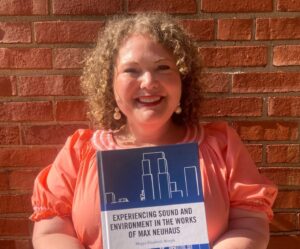 A smiling woman in a peach dress stands against a brick wall, holding a book titled Experiencing Sound and Environment in the Works of Max Neuhaus..