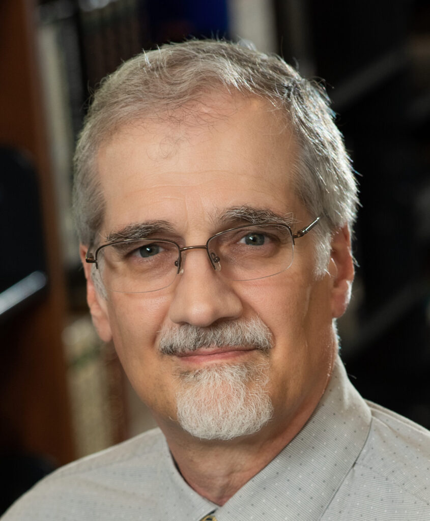 Portrait of an older man with gray hair, glasses, and a goatee, wearing a light gray collared shirt, looking at the camera with a slight smile. The background is softly blurred.
