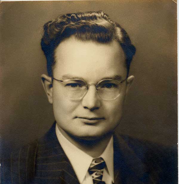 Black-and-white portrait of a man with glasses and wavy hair, wearing a suit, striped tie, and collared shirt, looking directly at the camera with a neutral expression.