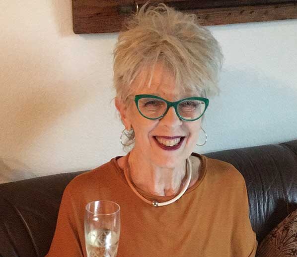 Smiling older woman with short gray hair, green glasses, and hoop earrings sits on a couch holding a glass of champagne. She wears an orange top and a silver necklace against a white wall background.