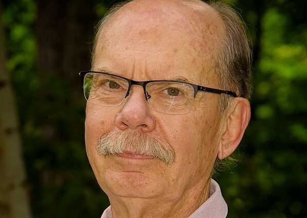 Older man with glasses, balding head, and a mustache, wearing a light-colored shirt, standing outdoors with a blurred background of green foliage.
