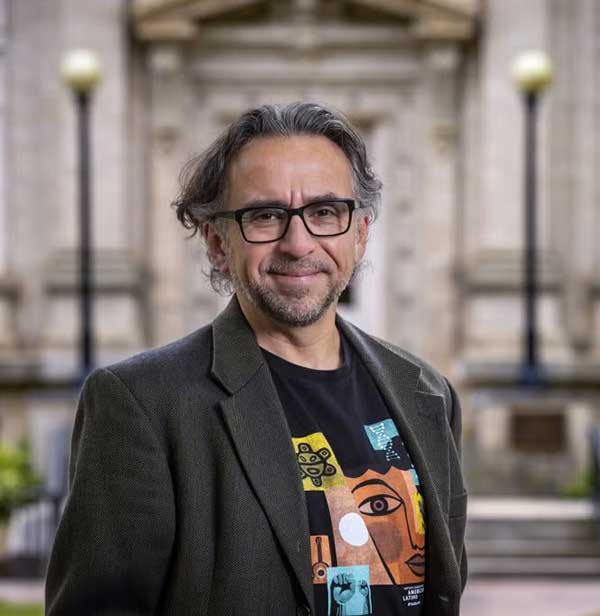 A man with glasses and graying hair smiles outdoors. He wears a dark blazer over a colorful graphic t-shirt and stands in front of a blurred stone building with lamp posts.