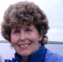 A woman with curly brown hair and a blue jacket smiles outdoors with water and sky in the background.