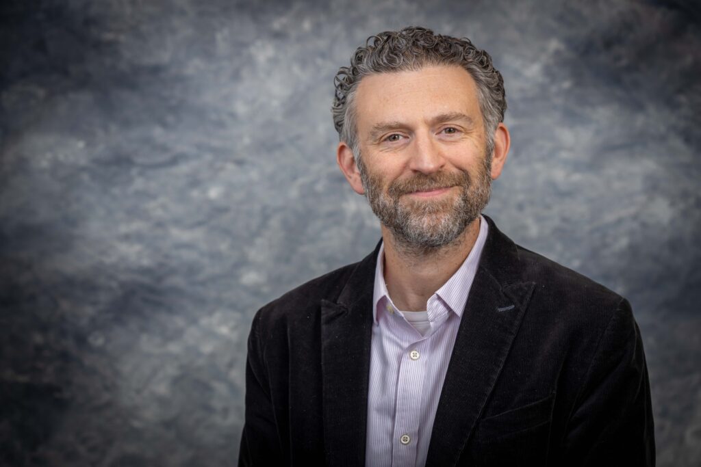 A man with curly hair and a beard, wearing a dark blazer and light shirt, smiles slightly while posing in front of a textured gray backdrop.