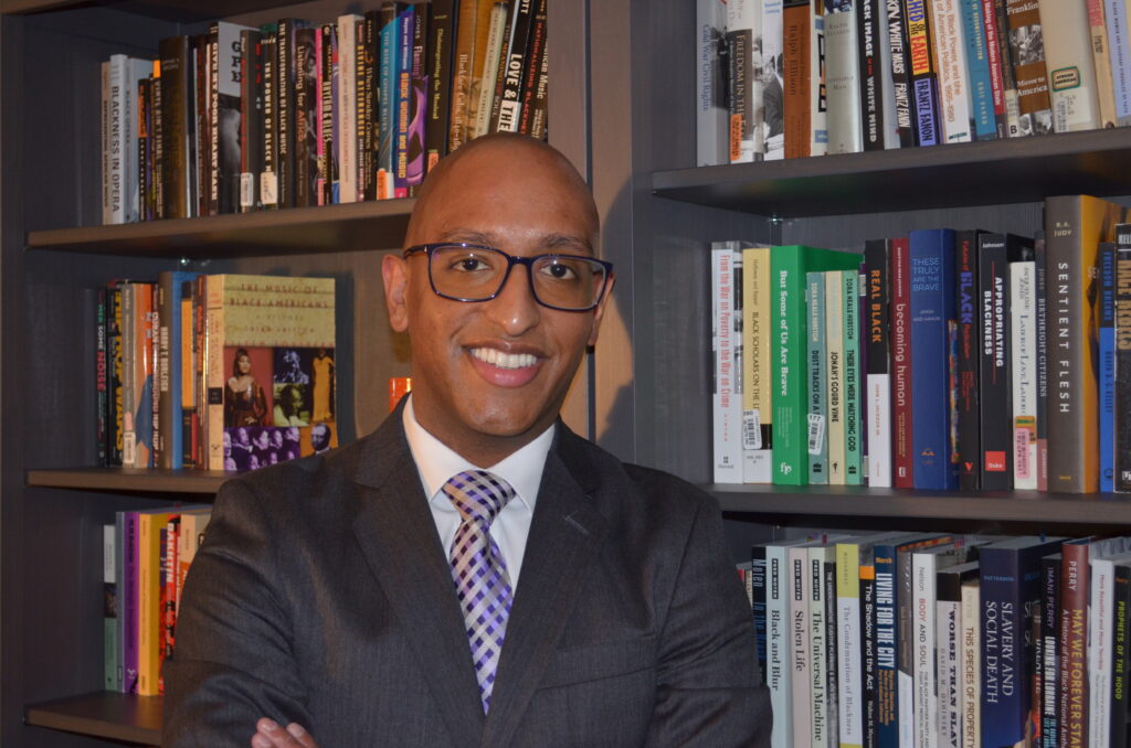 A person in a suit and glasses stands smiling with arms crossed in front of bookshelves filled with various books.