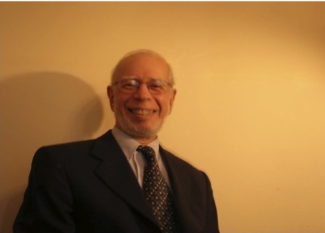 An older man in a suit and tie smiles at the camera, standing against a plain beige wall with soft lighting.