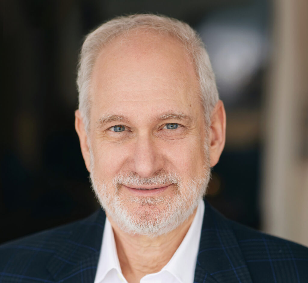 Smiling older man with short white hair and a trimmed white beard, wearing a dark plaid suit jacket and white shirt, posing in front of a blurred dark background.