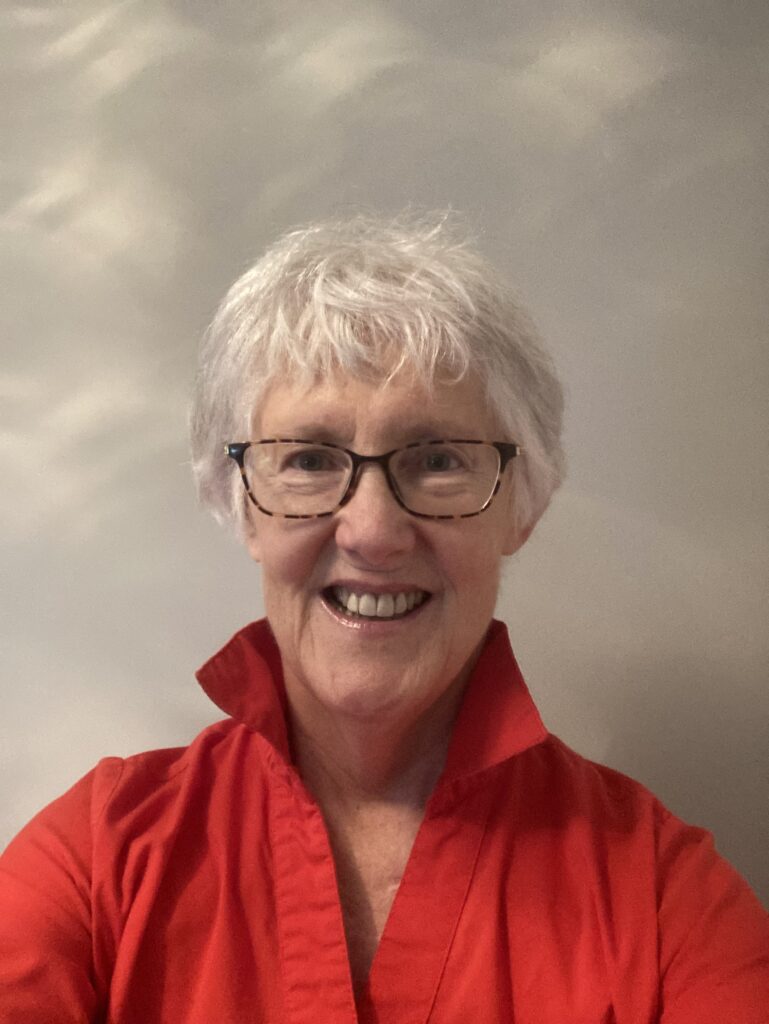 Older woman with short white hair and glasses smiles at the camera, wearing a bright red collared shirt against a neutral, softly lit background.