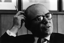 A man wearing glasses and a suit sits indoors, holding a pencil to his head and looking thoughtful. The background shows wood paneling and framed pictures. The photo is black and white.
