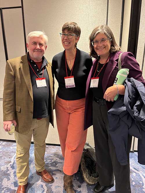 Three adults wearing conference badges stand together, smiling. The man on the left wears a tan blazer; the two women on the right wear glasses, with one in coral pants and the other holding a jacket and a green water bottle.