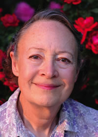 An older woman with short gray hair smiles gently. She is wearing a patterned blouse and is standing in front of blooming pink and red flowers.