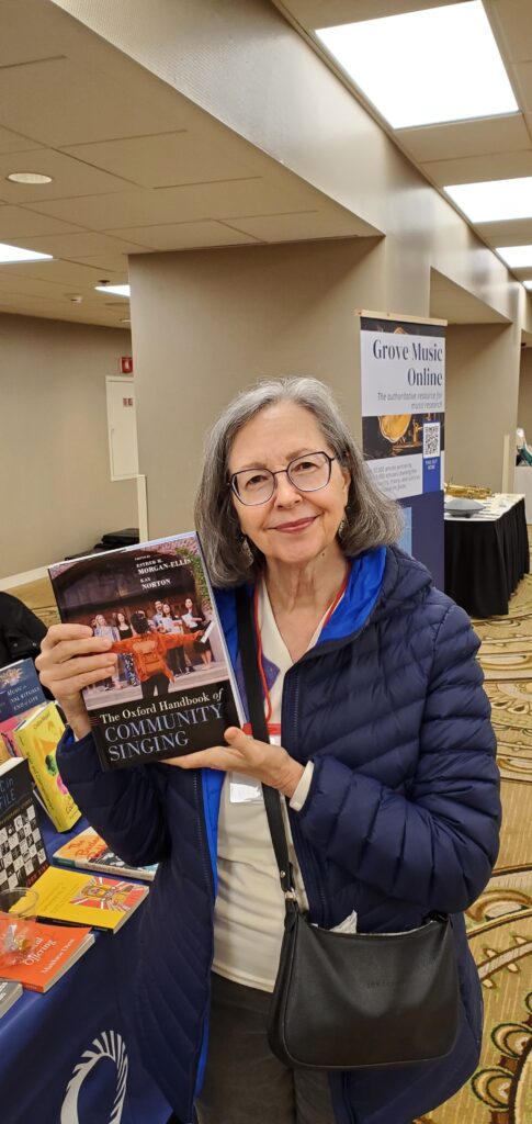 A woman with gray hair and glasses smiles while holding The Oxford Handbook of Community Singing at a conference book display.
