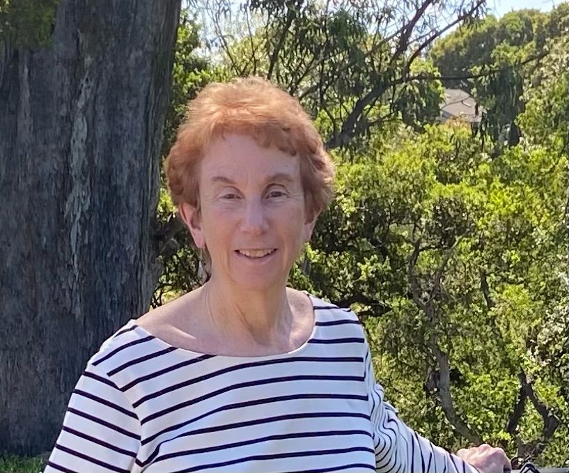 A woman with short, light brown hair wearing a white and black striped top stands outdoors in front of trees on a sunny day.