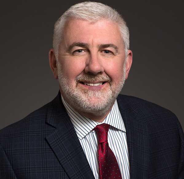 Smiling man with short gray hair and beard, wearing a dark plaid suit, white striped shirt, and red tie, posing against a plain dark background.
