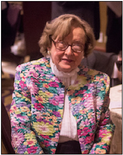 An elderly woman with glasses and short hair sits at a table, wearing a colorful floral jacket and a white turtleneck.
