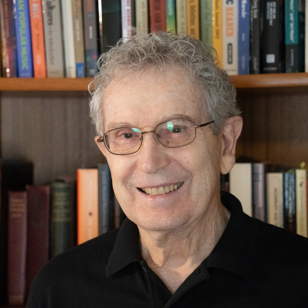 An older man with curly gray hair and glasses smiles while standing in front of a bookshelf filled with books. He is wearing a black collared shirt.