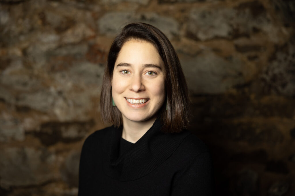 A woman with straight brown hair and a black sweater smiles in front of a stone wall background.