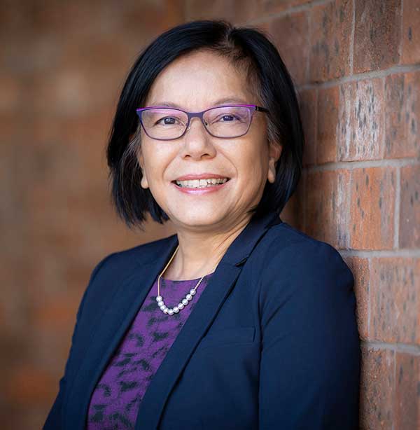A woman with short black hair, glasses, and a navy blazer over a purple top smiles while leaning against a brick wall.