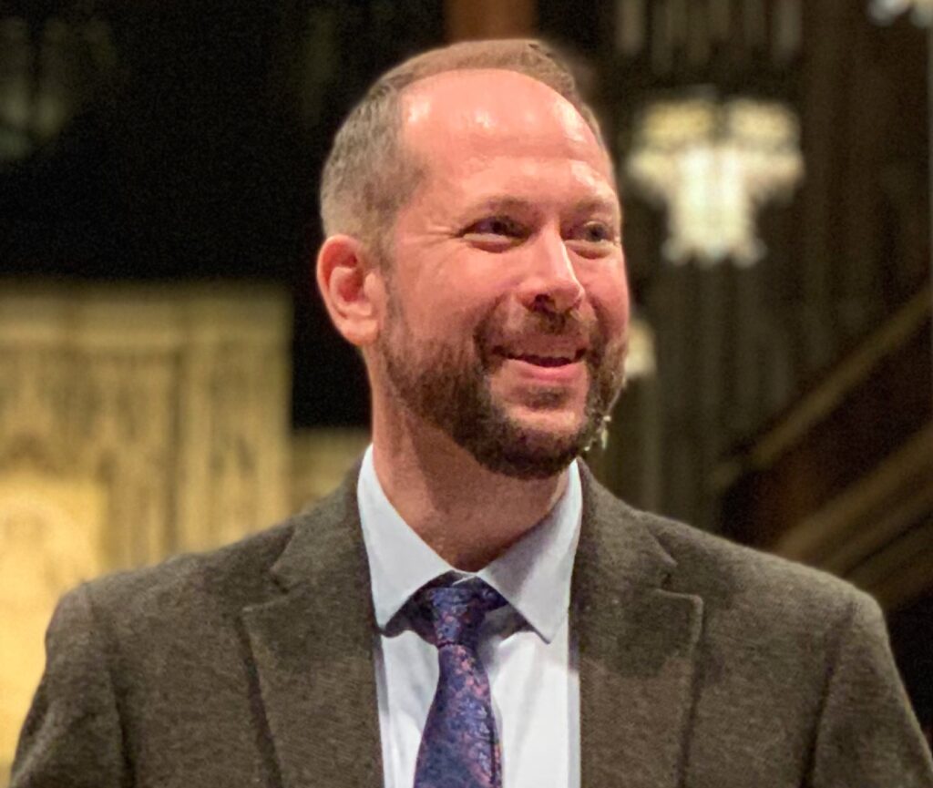 A man with short hair and a beard, wearing a suit jacket, dress shirt, and patterned tie, smiles while standing indoors with blurred architectural details in the background.
