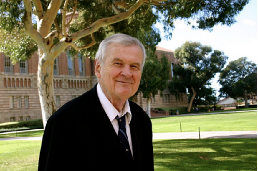 An older man in a suit and tie stands outdoors on a sunny day with trees, grass, and a large brick building in the background.