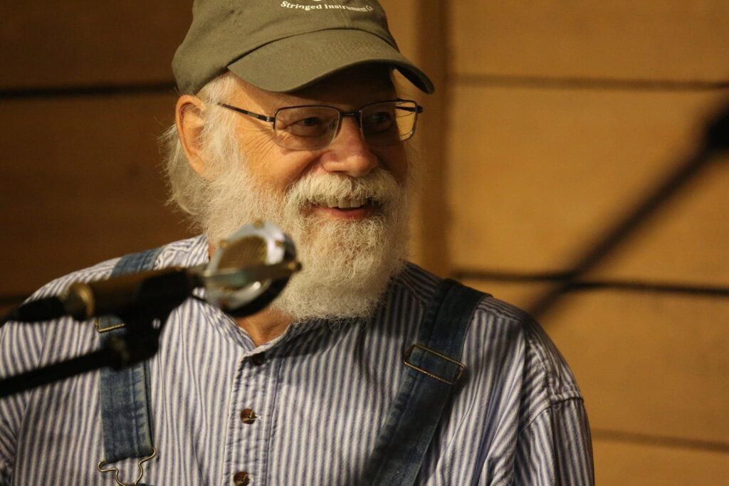 An older man with a white beard, glasses, and a cap smiles while sitting at a microphone, wearing a striped shirt and overalls against a wooden background.