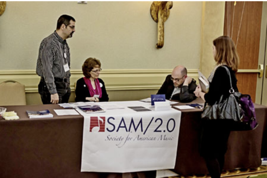 Four people interact at a conference table labeled SAM/2.0 Society for American Music, with pamphlets and materials displayed on the table in a hotel meeting room.