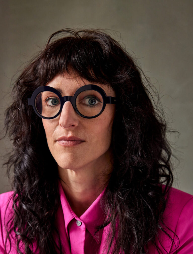 A person with long, wavy dark hair and large round black glasses wears a bright pink shirt and looks at the camera against a neutral background.