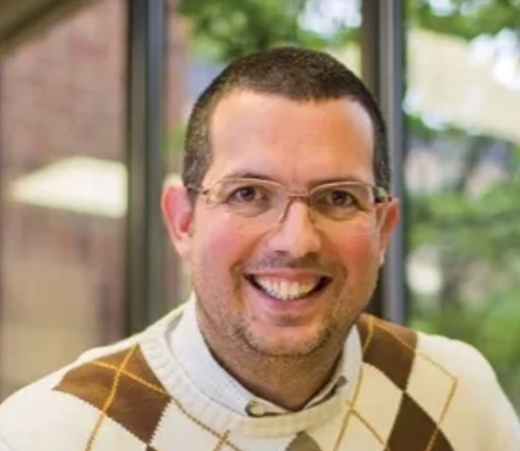 A smiling man with short dark hair and glasses, wearing a white sweater with brown argyle pattern, stands indoors with large windows and greenery visible outside.