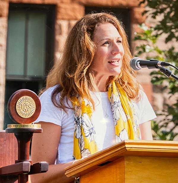 A woman with long light brown hair speaks at a podium outdoors. She wears a white shirt and yellow floral scarf. A microphone and a wooden emblem are visible. Sunlight illuminates her face and background.
