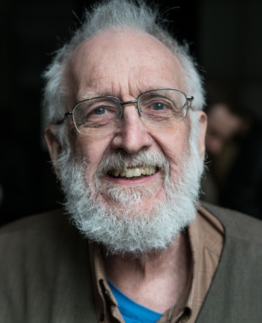 An elderly man with glasses and a white beard smiles warmly at the camera, wearing a brown jacket over a blue shirt. The background is softly blurred.