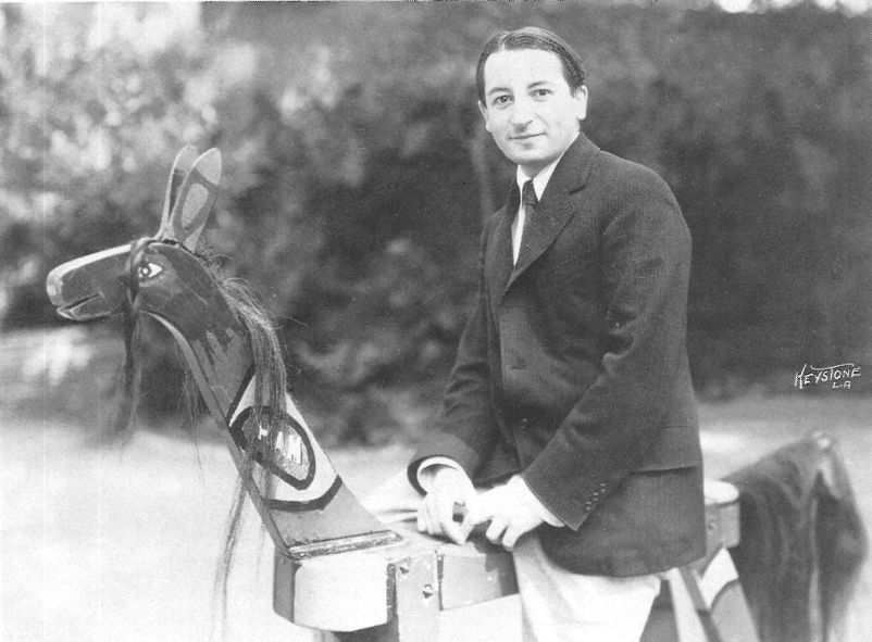 A man in a suit sits on a painted wooden horse outdoors, smiling at the camera. The background is blurred greenery.