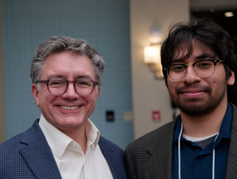 Two people wearing glasses and suits stand together indoors, smiling at the camera. The background is softly lit with a wall lamp visible behind them.