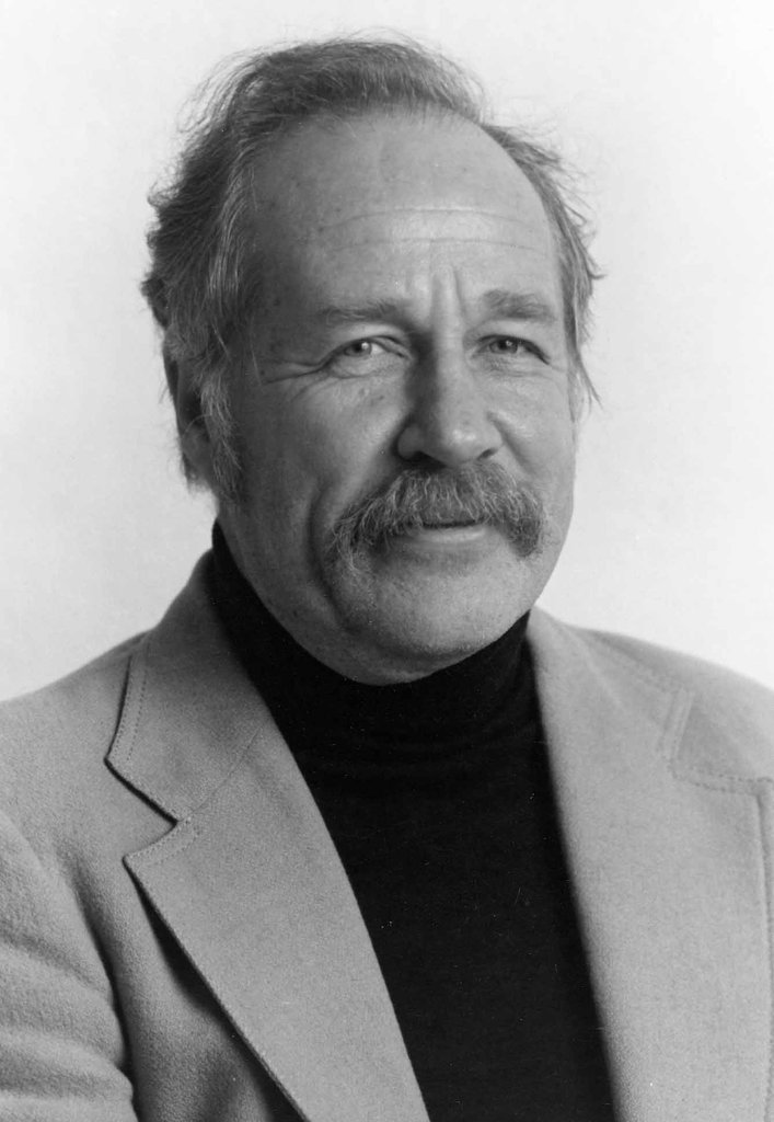 A man with a mustache and light-colored hair wearing a light blazer over a dark turtleneck, posing in front of a plain light background.