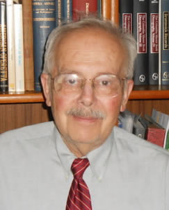 An older man with glasses, a gray mustache, and short hair, wearing a light shirt and red striped tie, sits in front of a bookshelf filled with books.