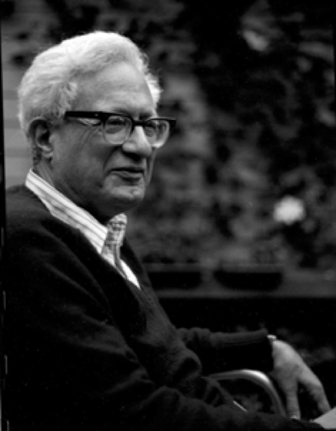 An older man with white hair and glasses sits outdoors, wearing a dark sweater over a collared shirt. He faces slightly to the side and smiles gently, with a blurred leafy background behind him.