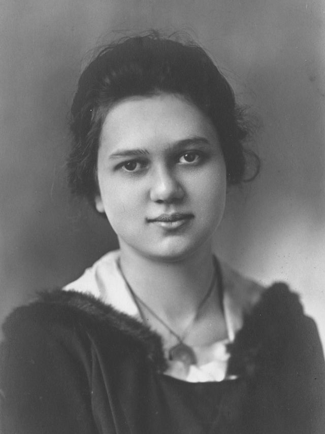Black and white portrait of a young woman with dark hair pulled back, wearing a dark top with a light collar, looking slightly past the camera and softly smiling.
