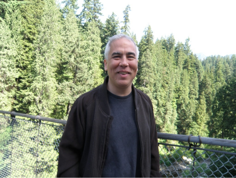 A man with gray hair, wearing a dark jacket and shirt, stands on a bridge with a metal railing, smiling. Tall green trees and a forest landscape fill the background under a bright sky.