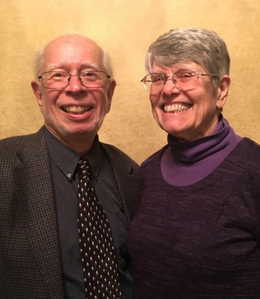 An older man in a suit and tie stands smiling next to an older woman wearing glasses and a purple sweater. Both are smiling and posing in front of a beige wall.