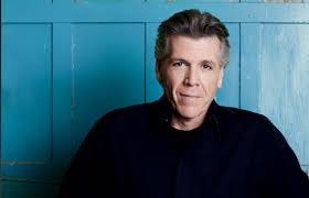 A middle-aged man with short gray hair wearing a black shirt sits in front of a blue wooden wall, looking at the camera with a neutral expression.