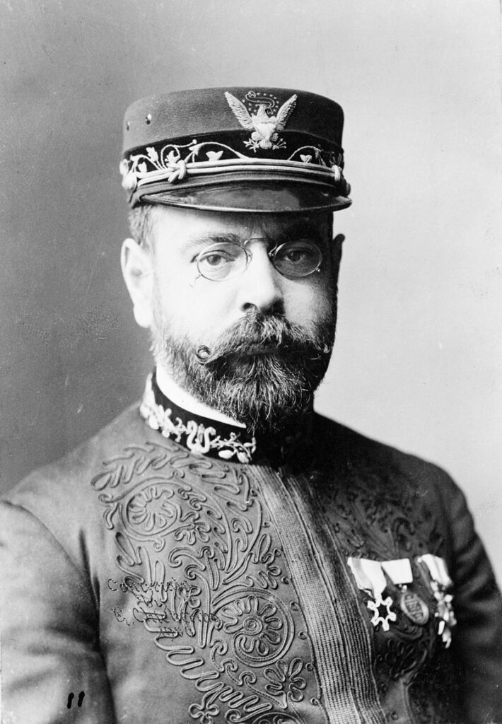 Black and white portrait of a man with glasses, a mustache, and beard, wearing a decorated military-style uniform and hat with an eagle emblem, and medals pinned to his chest.