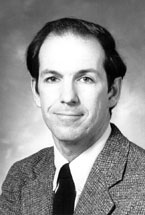 Black and white portrait of a man in a suit and tie, with short dark hair, looking at the camera and smiling slightly, set against a plain background.