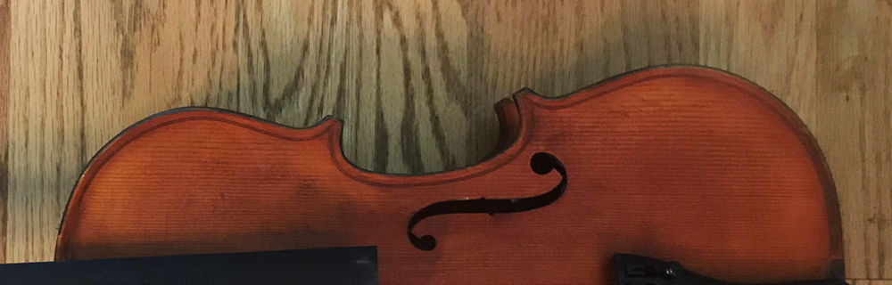 A close-up of the upper half of a violin resting on a wood floor, showing part of the f-hole and the violins reddish-brown body.