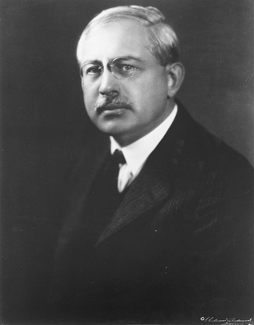 Black-and-white portrait of an older man with glasses, a mustache, and short hair, wearing a suit and tie, looking slightly to the side against a dark background.