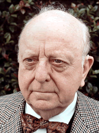 An elderly man with a serious expression, bald head, and wearing a checkered jacket and bow tie stands outdoors in front of green foliage.